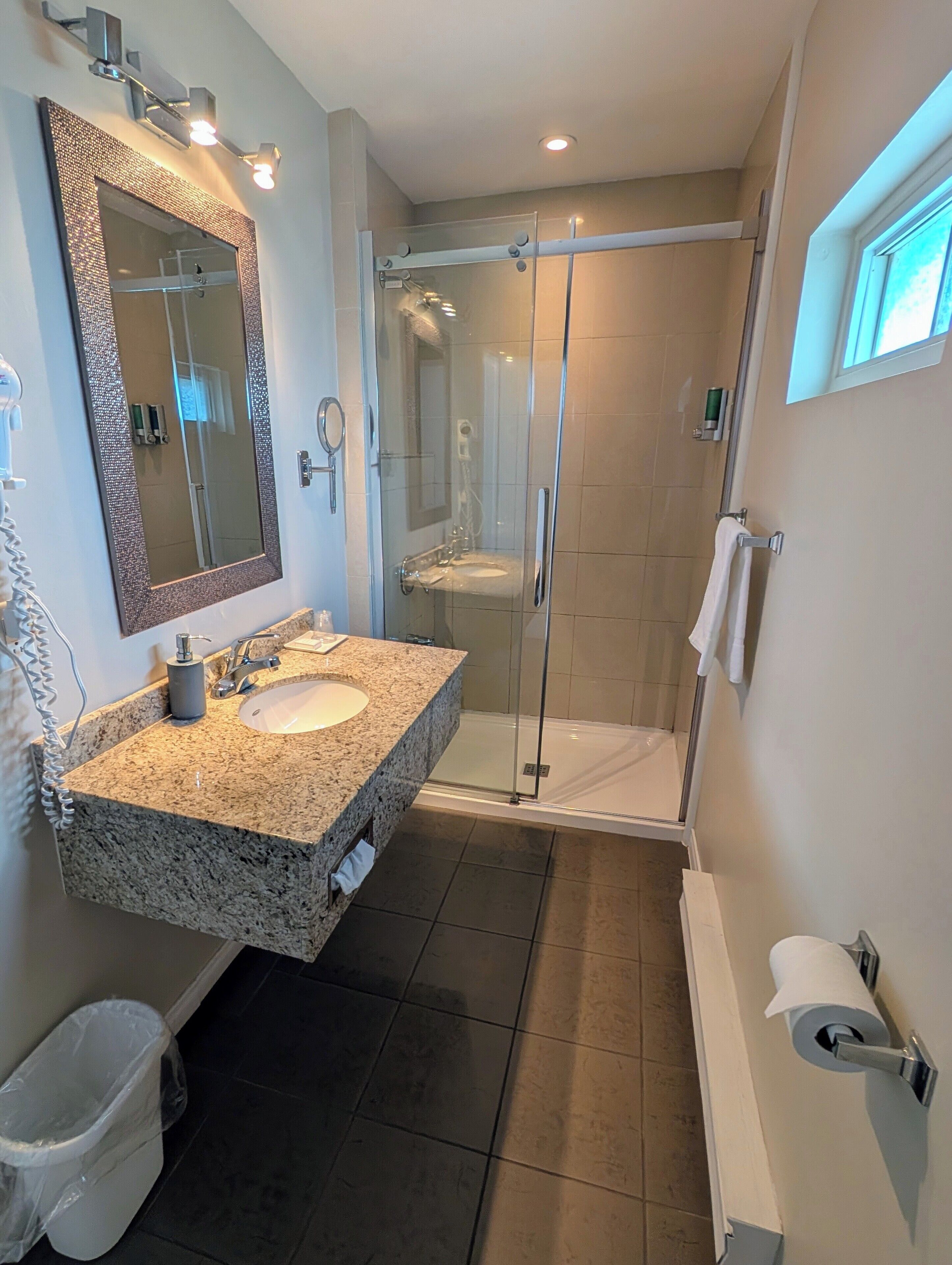 A bathroom with a granite sink, a large mirror, a glass shower enclosure, and a toilet paper holder.