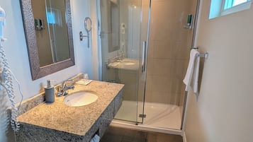 A bathroom with a granite sink, a large mirror, a glass shower enclosure, and a toilet paper holder.