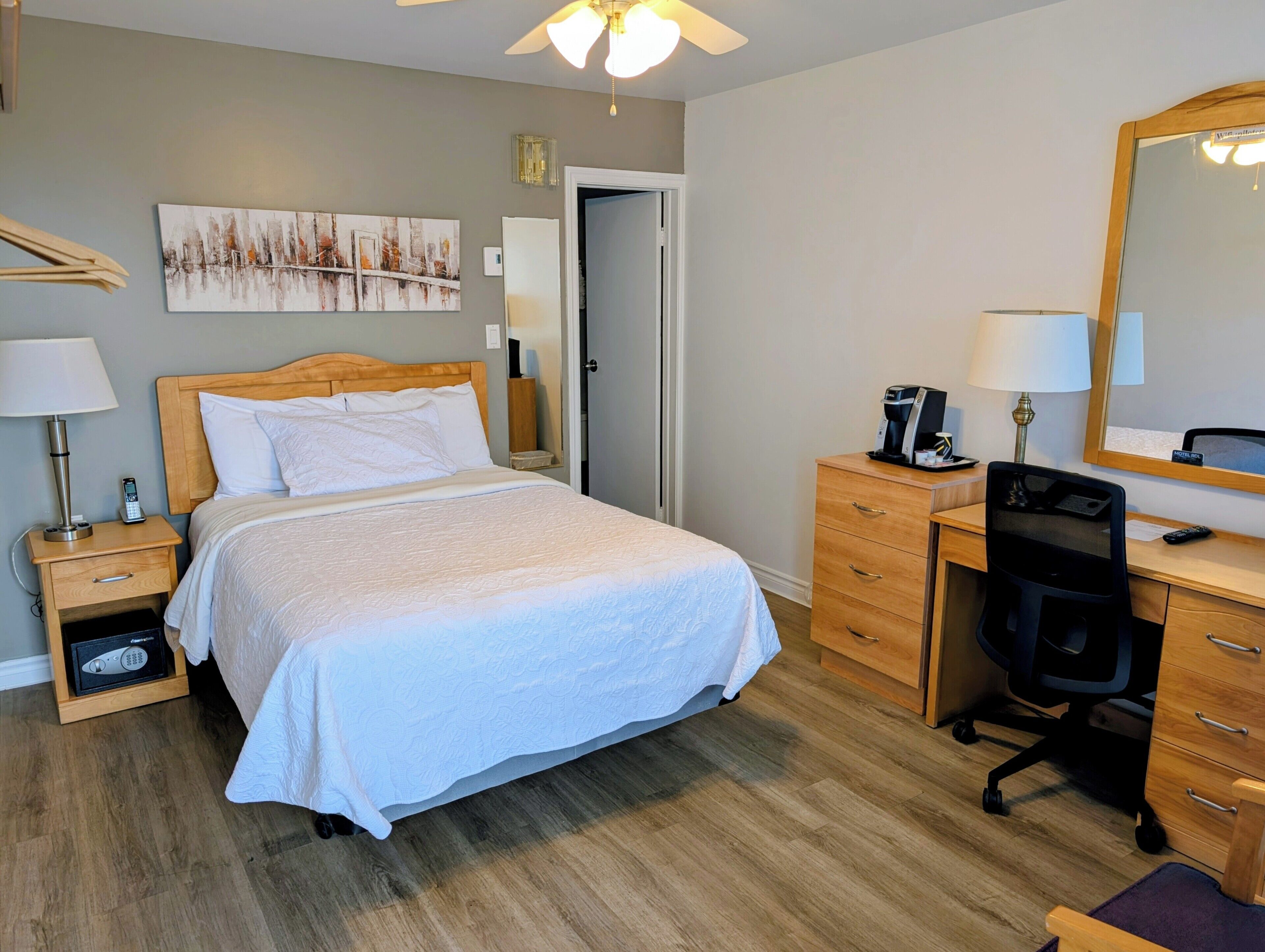 A hotel room with a bed, a desk, a chair, a mirror, a painting, and a ceiling fan.