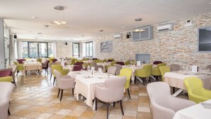 A spacious dining area with multiple tables and chairs, large windows, and a brick wall. - Albergo Lenno (Tremezzina)