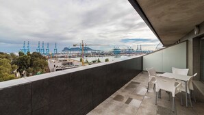 Un balcón con vista a un puerto, incluyendo grúas y barcos, y una mesa con sillas.