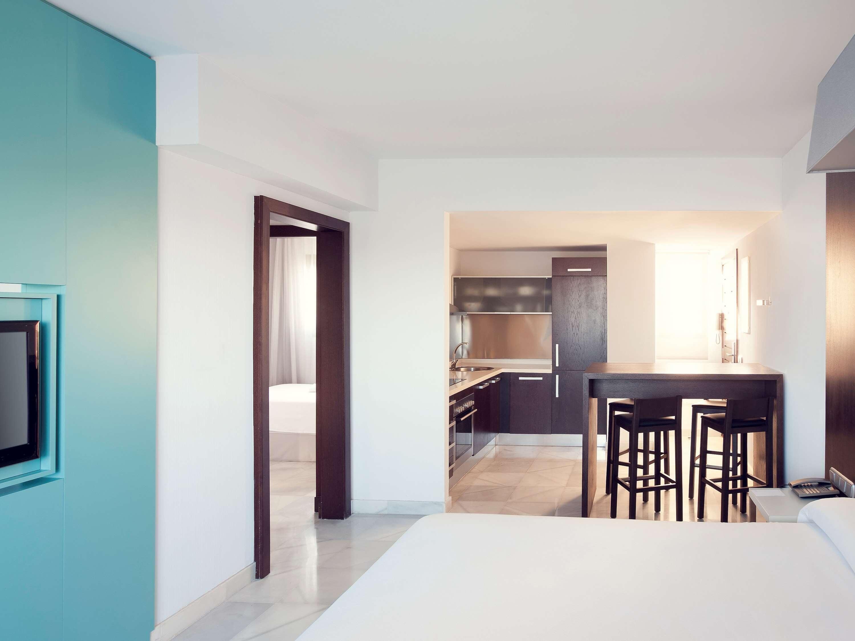 A modern hotel room with a bed, a desk, and a kitchenette.