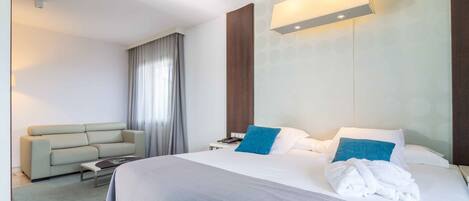 Una habitación de hotel moderna con una cama grande, un sofá, una mesita de noche y un ventanal con cortinas.