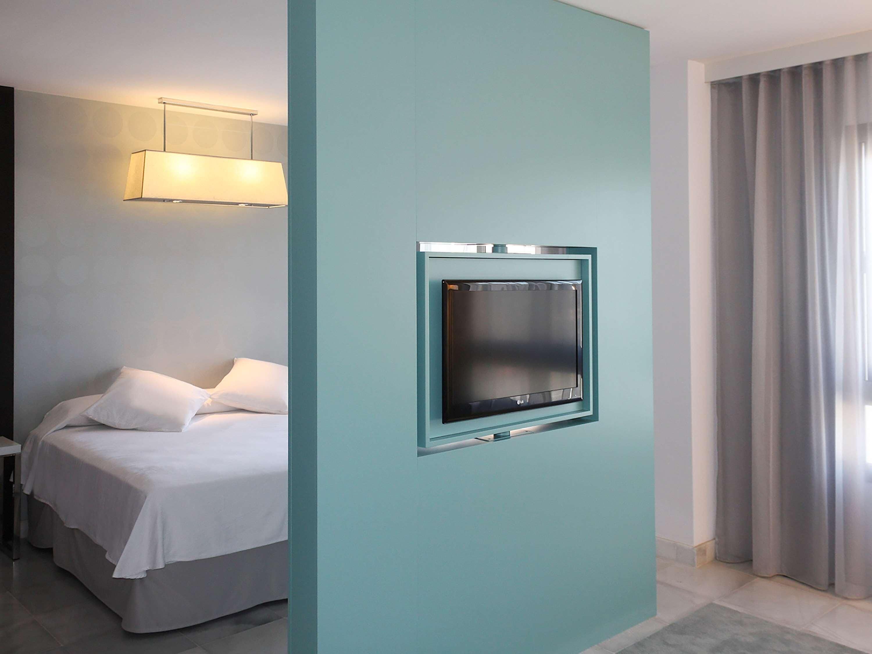 A hotel room with a bed, a flat-screen TV mounted on a teal-colored wall, a bedside table, and a window with curtains.