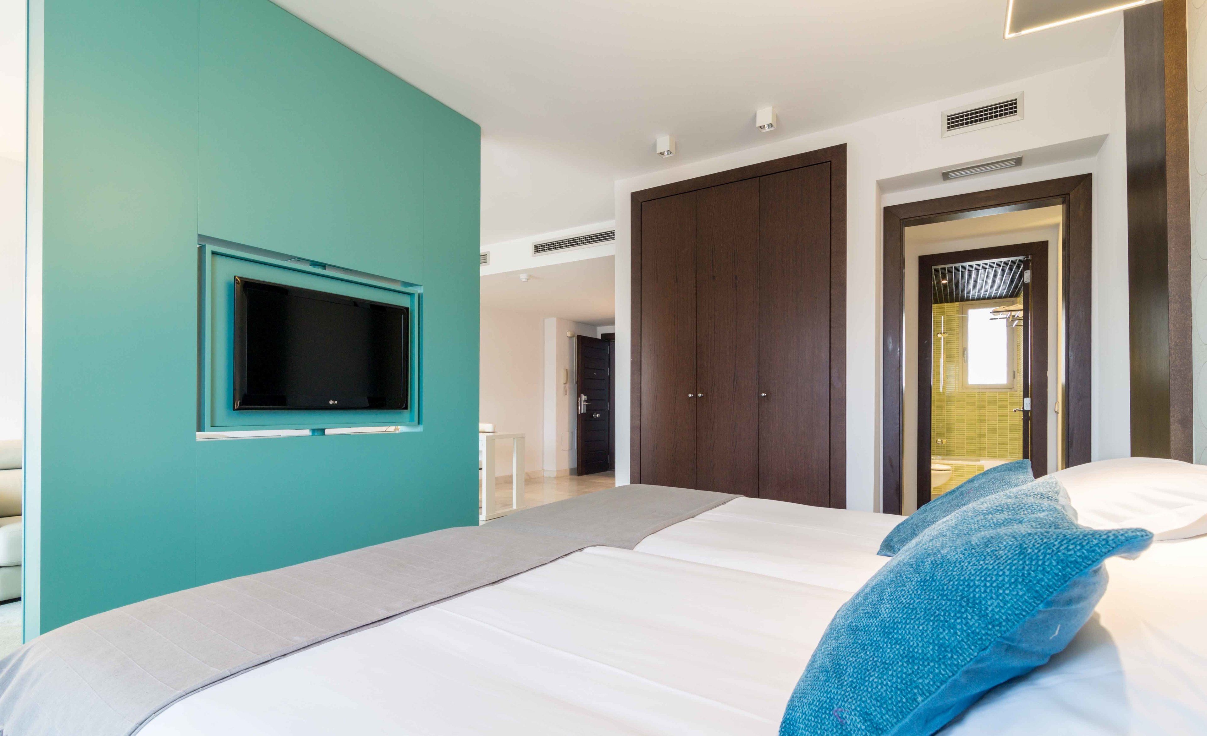 Un dormitorio moderno con una pared de acento color turquesa, una cama grande con ropa de cama blanca, un televisor de pantalla plana montado en la pared y un armario con puertas de madera oscura.