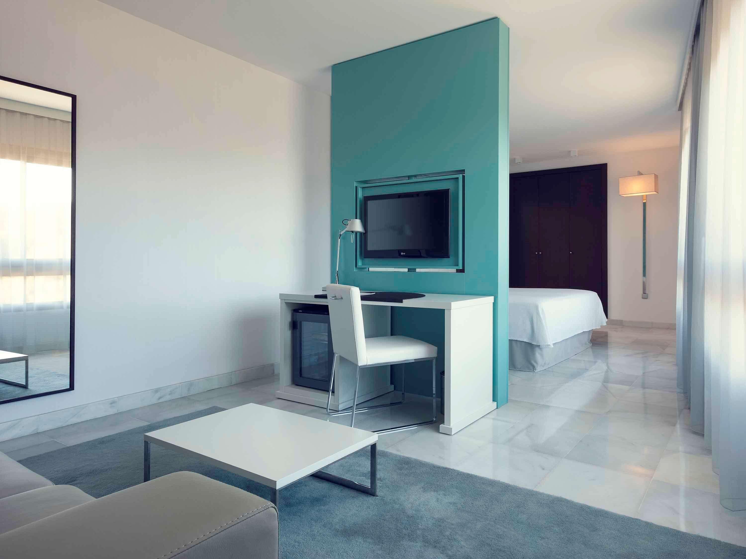 A modern hotel room with a teal accent wall, a flat-screen TV, a desk with a chair, a bed, and a lamp.