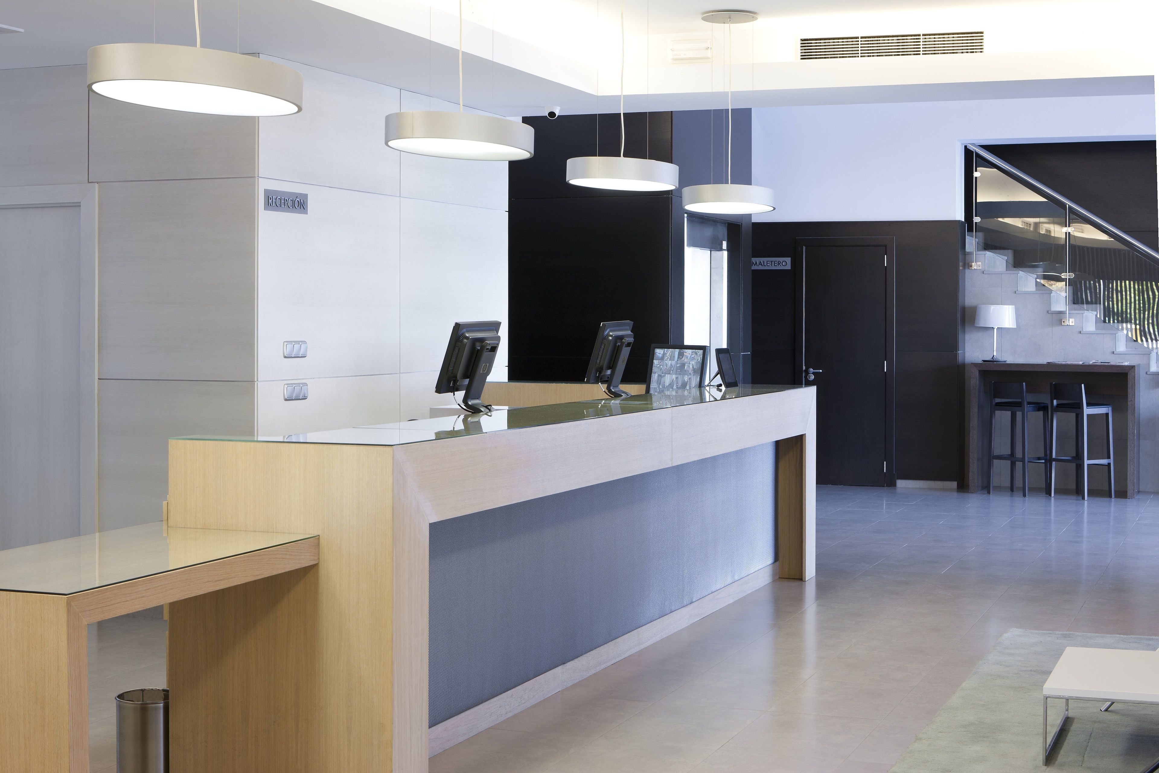 A modern reception desk with a glass top, overhead lighting, and a staircase leading to an upper level.