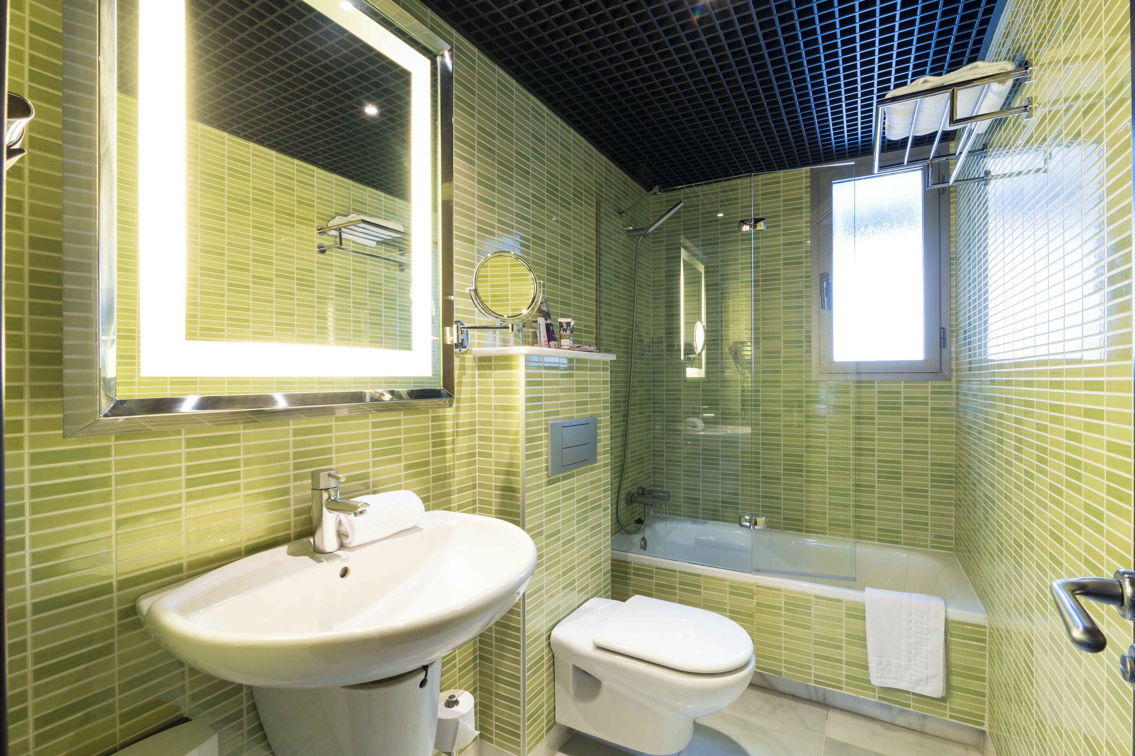 Een badkamer met een wastafel, toilet en bad, allemaal met groene betegelde wanden en een plafondrooster.