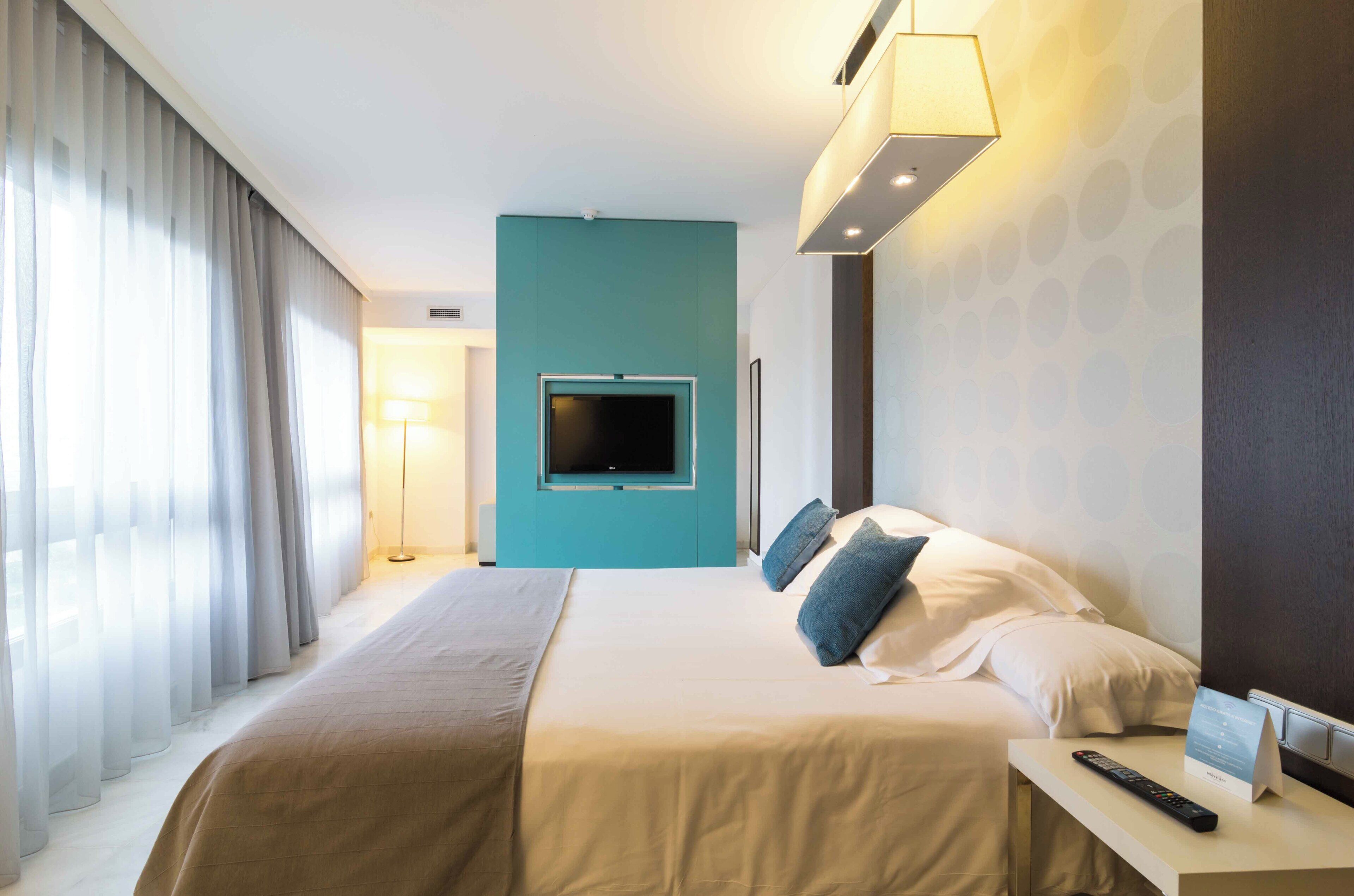Habitación de hotel con cama, televisor montado en la pared, mesita de noche con control remoto y lámpara.