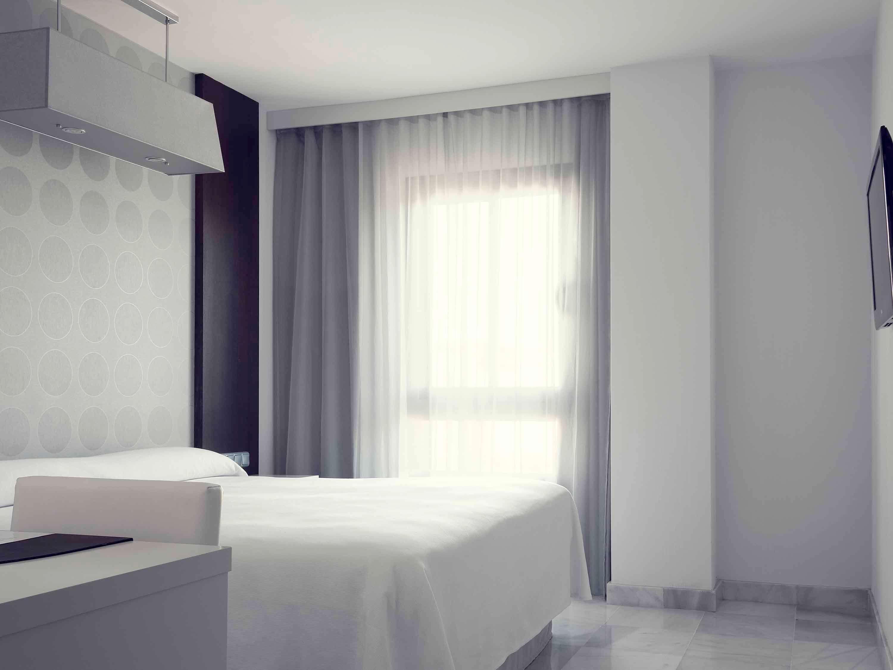 A modern hotel room with a bed, a bedside table, and a large window with curtains.