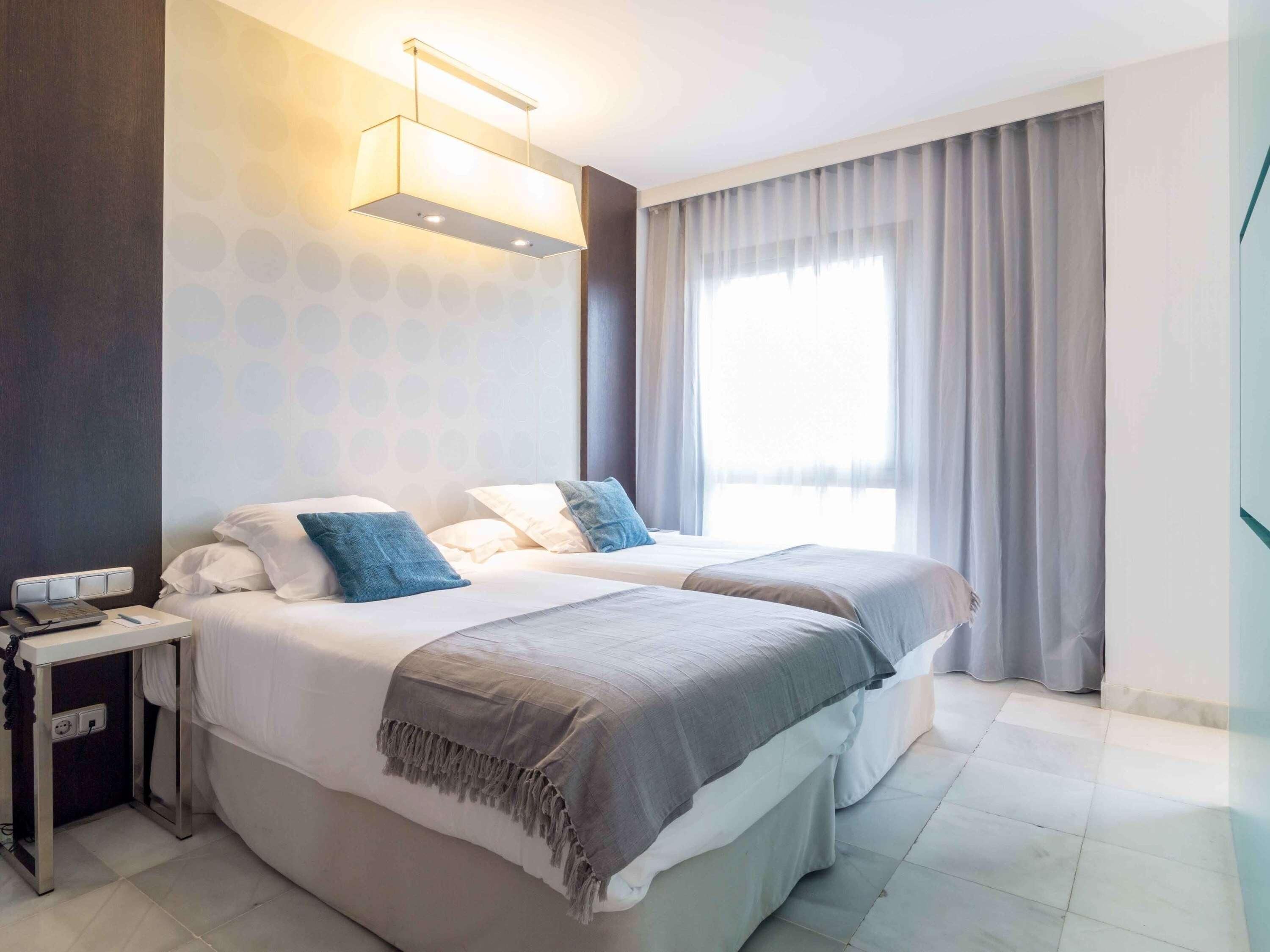 A modern hotel room with two beds, a bedside table, a lamp, and a large window with curtains.