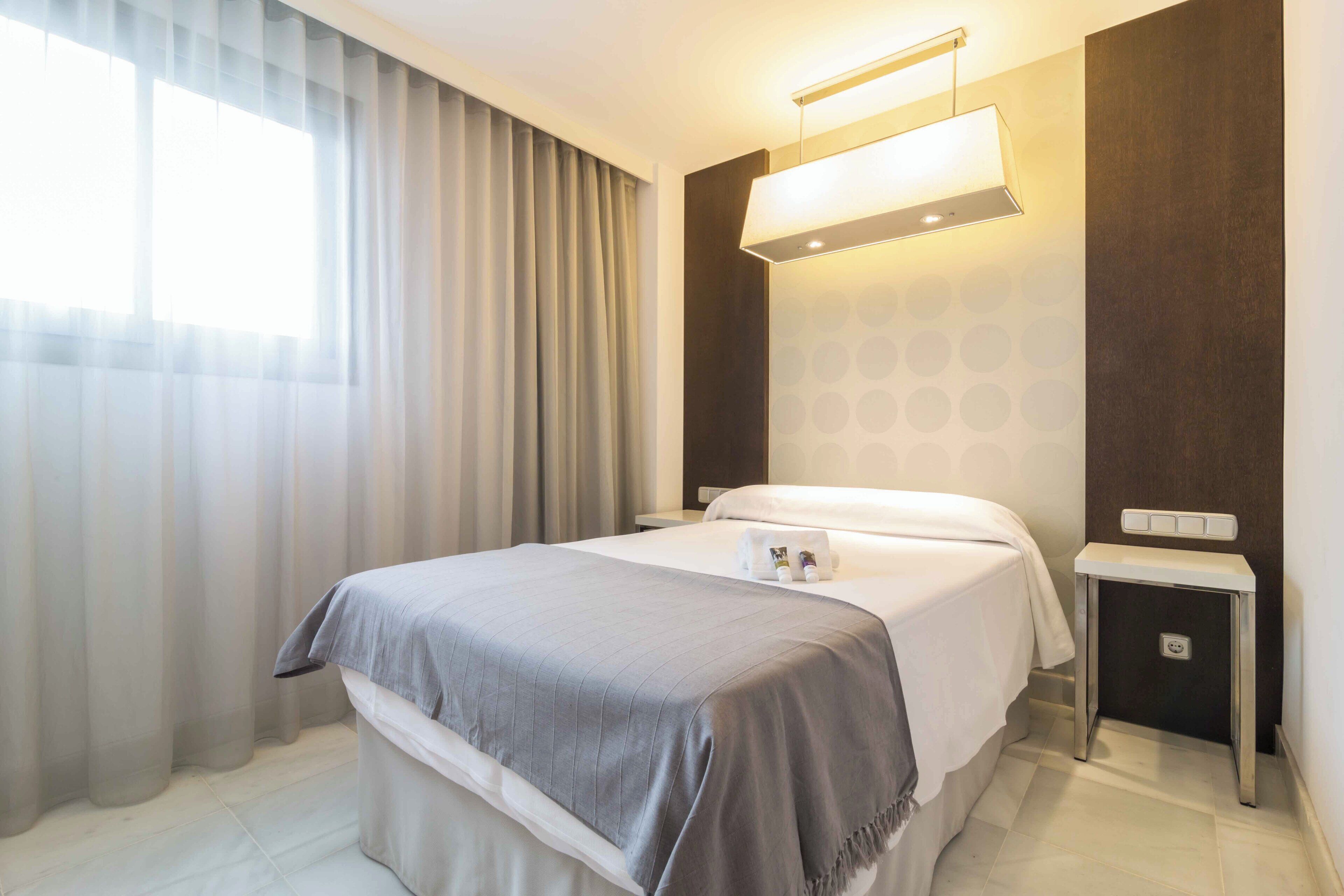 A hotel room with a bed, a bedside table, a nightstand, a lamp, and a window with curtains.