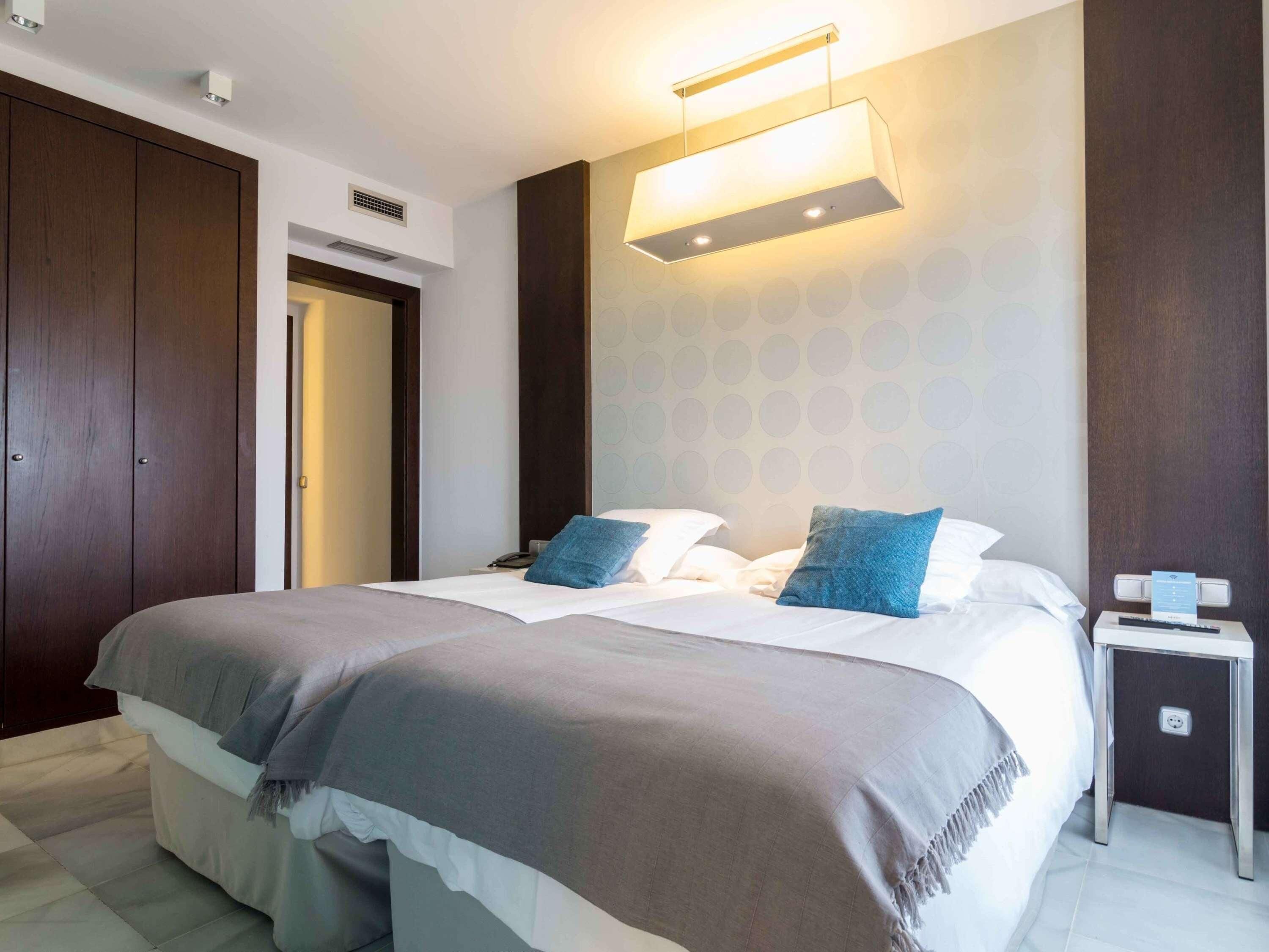 A modern hotel room with a large bed, two pillows, a bedside table, and a wall-mounted light fixture.