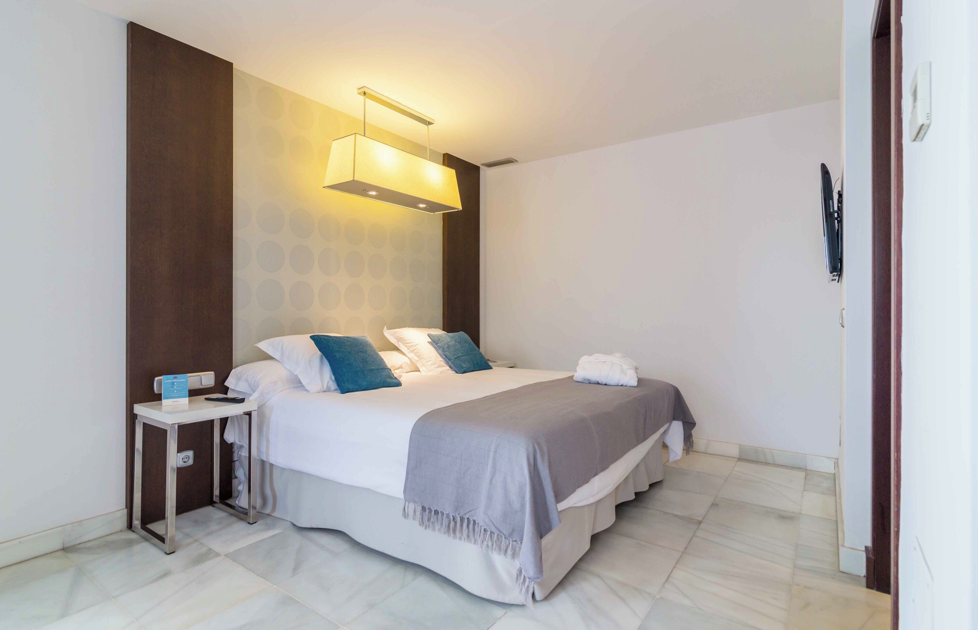 A modern hotel room with a large bed, a bedside table, a wall-mounted light, and a visible power outlet.