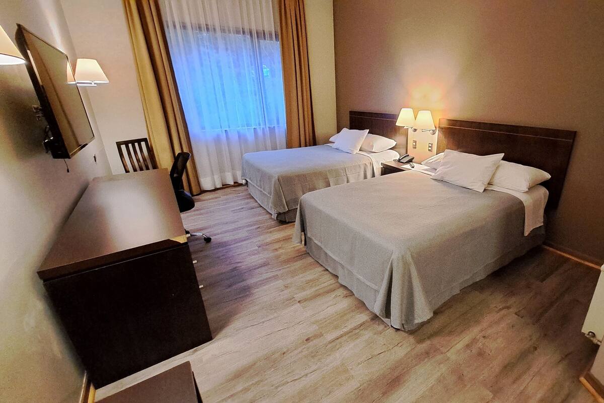 Une chambre d’hôtel avec deux lits, un bureau, une chaise, une télévision et une fenêtre avec des rideaux.