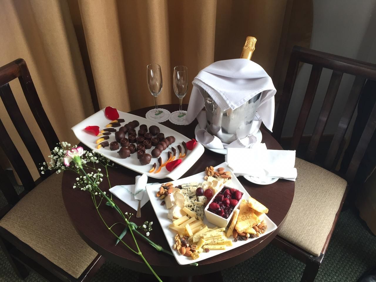 A round dining table with a variety of food items including chocolates, cheeses, fruits, and nuts. There are two wine glasses and a bottle of champagne on the table.