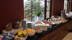 Disposición de un bufé con diversas opciones de comida, como frutas, postres y bebidas, dispuestas sobre un mostrador con vistas a zonas verdes al exterior.