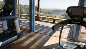 A treadmill and a stationary bike in a room with a wooden deck and a bench, overlooking a scenic landscape.