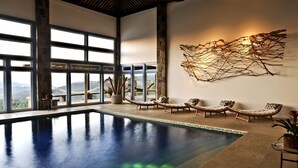 A spacious indoor swimming pool with clear views of the outdoors, a large wall-mounted art piece, and comfortable lounge chairs.
