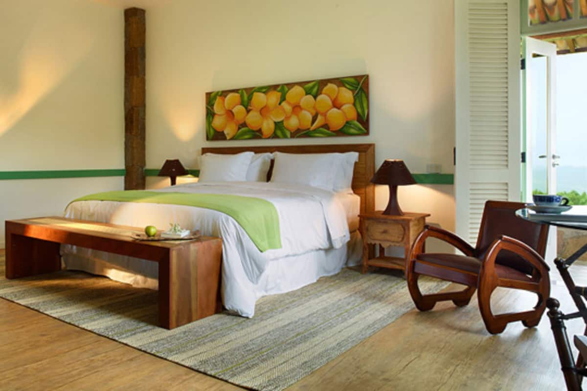 Een slaapkamer met een bed, nachtkastjes, een stoel, een houten bankje, een schilderij en een raam.