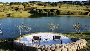 A circular stone seating area with two chairs and a small table, overlooking a lake with trees and a hilly landscape in the background.