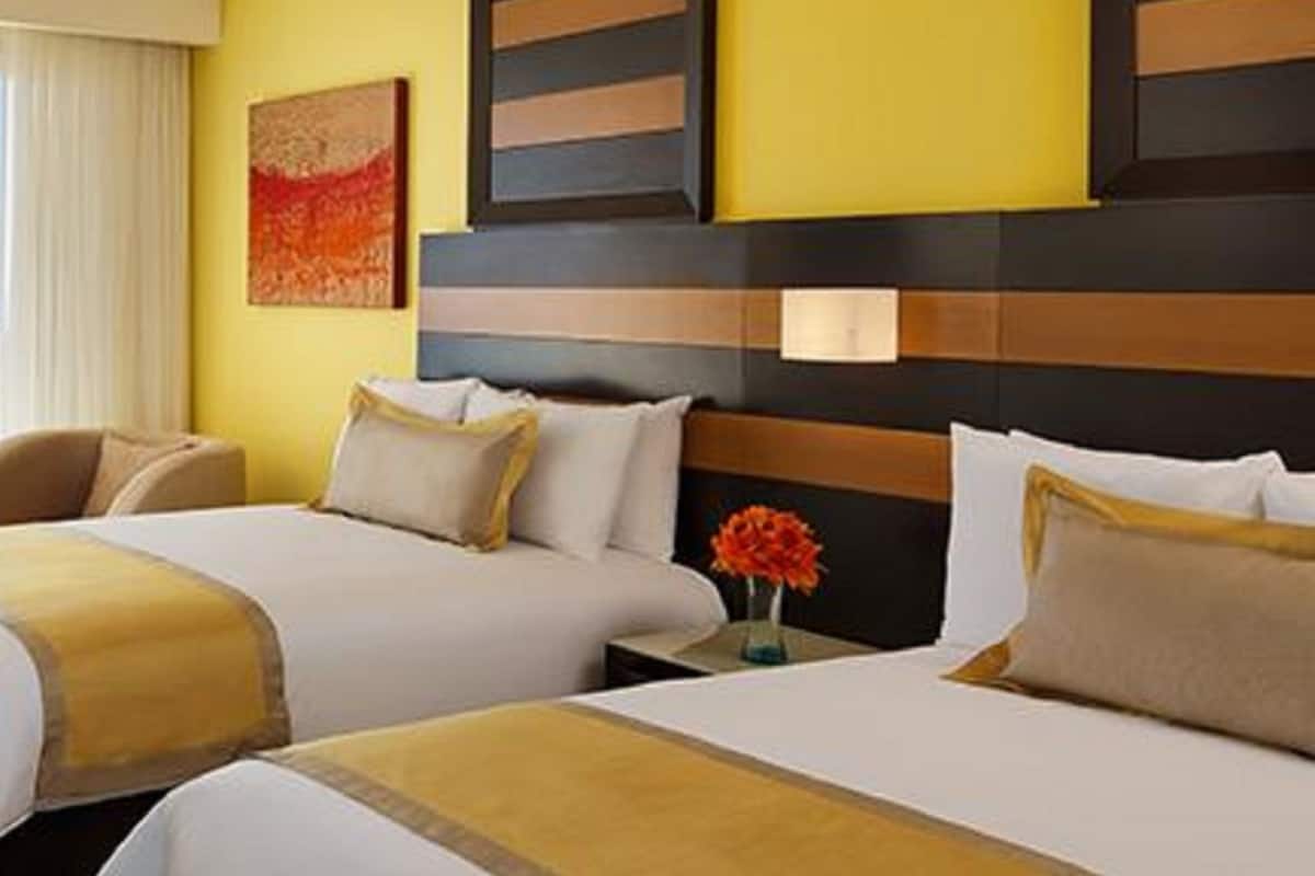 Ein Hotelzimmer mit zwei Betten, einem großen Fenster mit Vorhängen, einem Gemälde an der Wand und einer Vase mit Blumen auf dem Nachttisch.