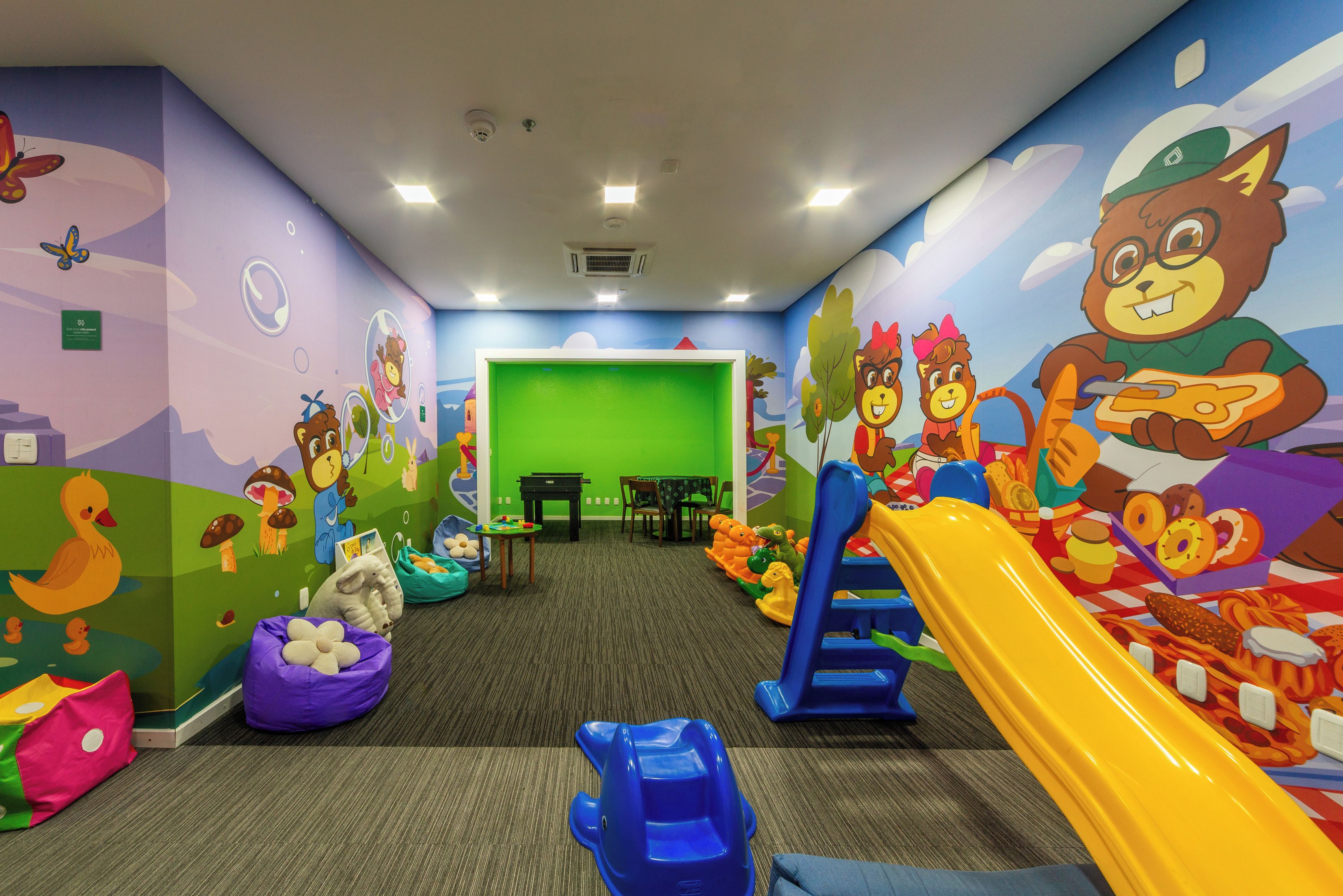 A children's play area with colorful murals, a slide, bean bags, and a green screen in the background.