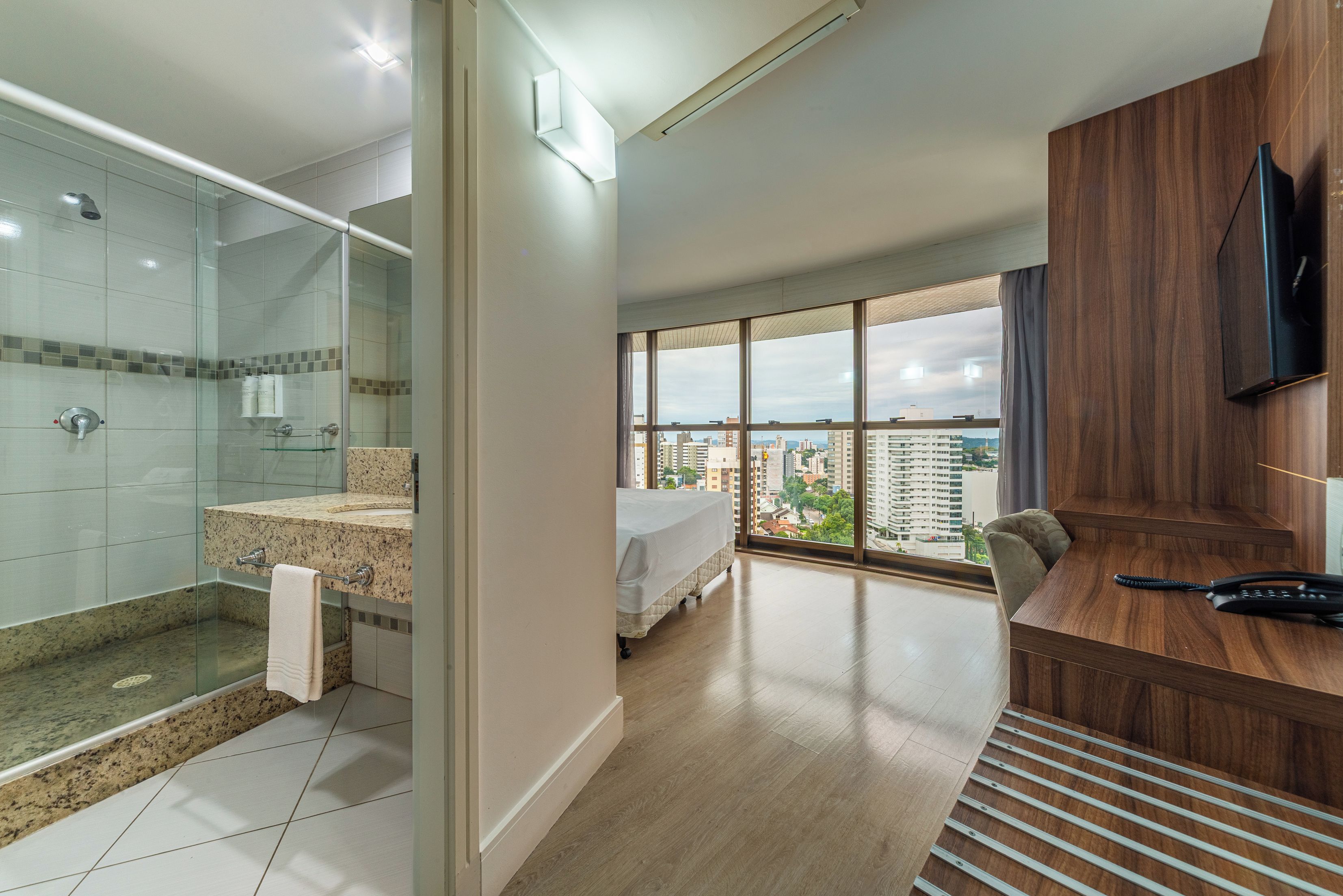 A modern hotel room with a glass-enclosed shower, a bed with a white mattress, a wooden desk with a telephone, and a large window offering a city view.