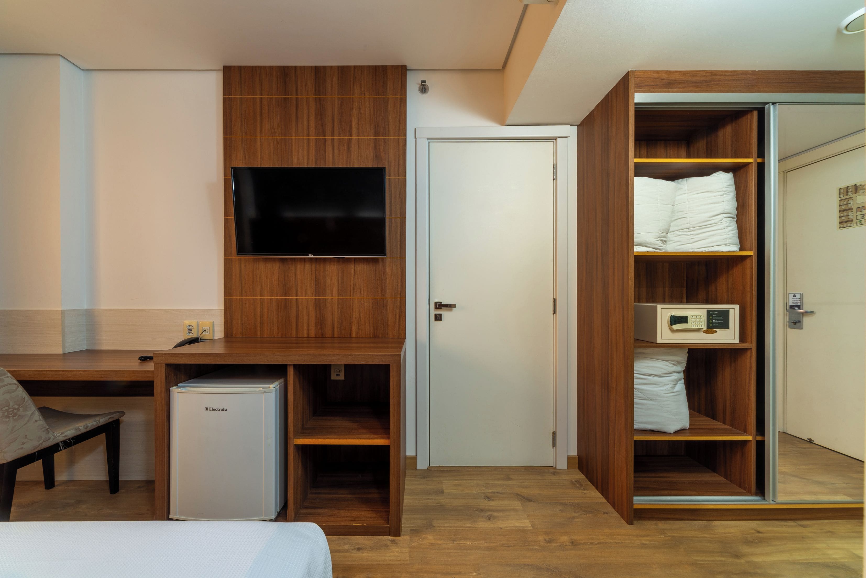 A compact hotel room with a wooden desk, a television, a mini-fridge, a wardrobe with a mirror, and a bed.