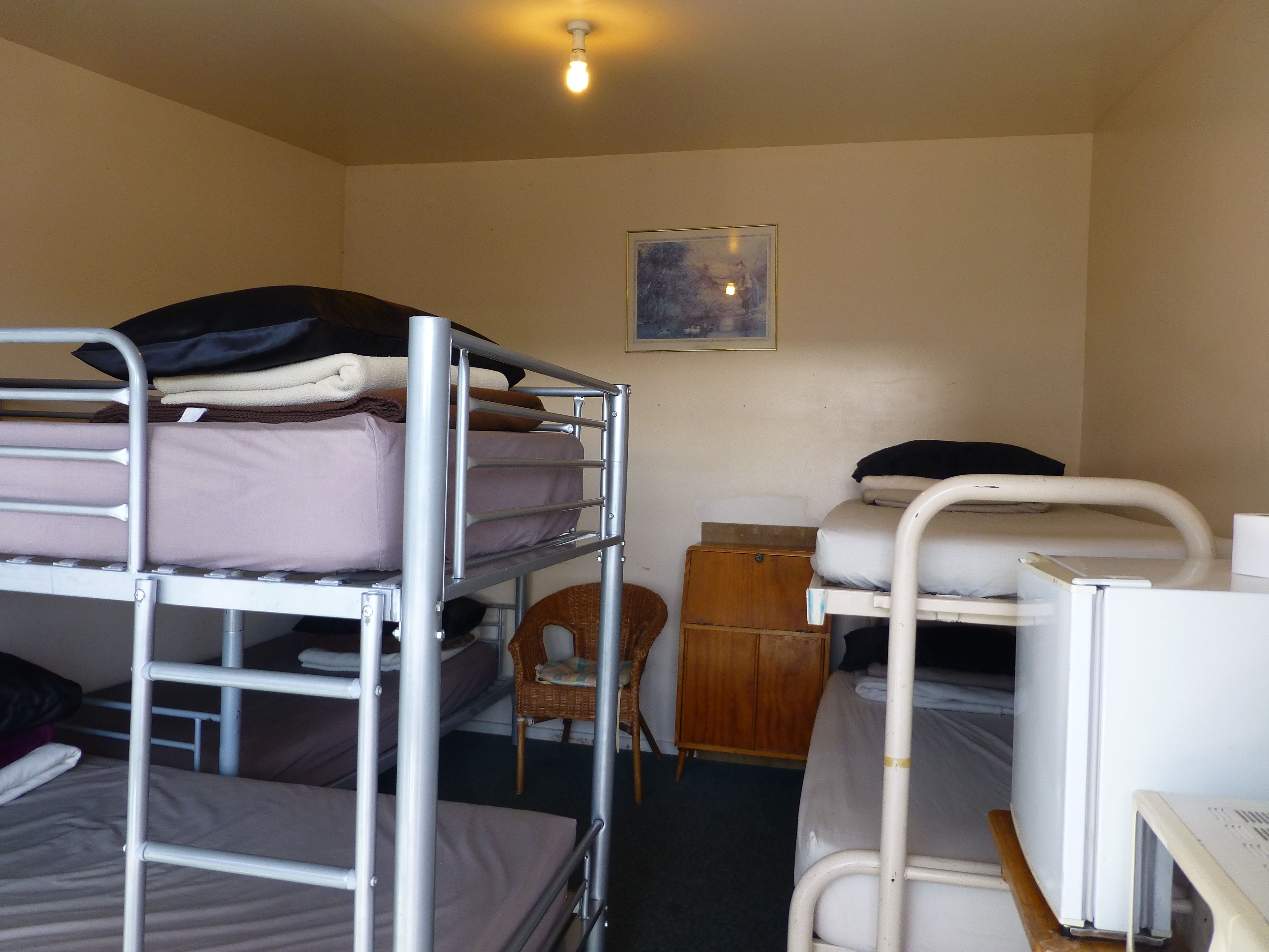 Ein Schlafsaalzimmer mit Etagenbetten, einem kleinen Holzschrank, einem Korbstuhl und einem gerahmten Bild an der Wand.