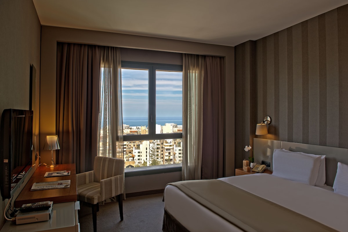 A hotel room with a large bed, a desk with a lamp, a chair, a nightstand with a phone, and a view of the city through the window.