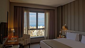 Una habitación de hotel con cama, escritorio con lámpara, silla, mesita de noche con teléfono y vista a la ciudad a través de amplios ventanales.