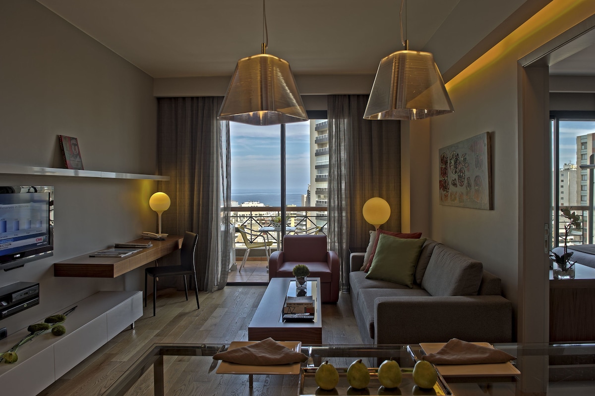 A modern living room with a sofa, coffee table, and TV. There is a balcony with a view of the city.
