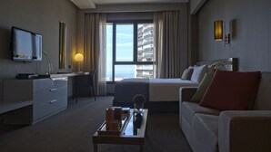 A hotel room with a bed, a desk with a TV, a coffee table, and a view of the city. - ParkTower Suites (Beirut)