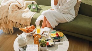 A woman in a white bathrobe sitting on a green sofa with a breakfast tray in front of her, including croissants, fruit, and a book titled "Face a Thymus-Boosting". - ParkTower Suites (Beirut)