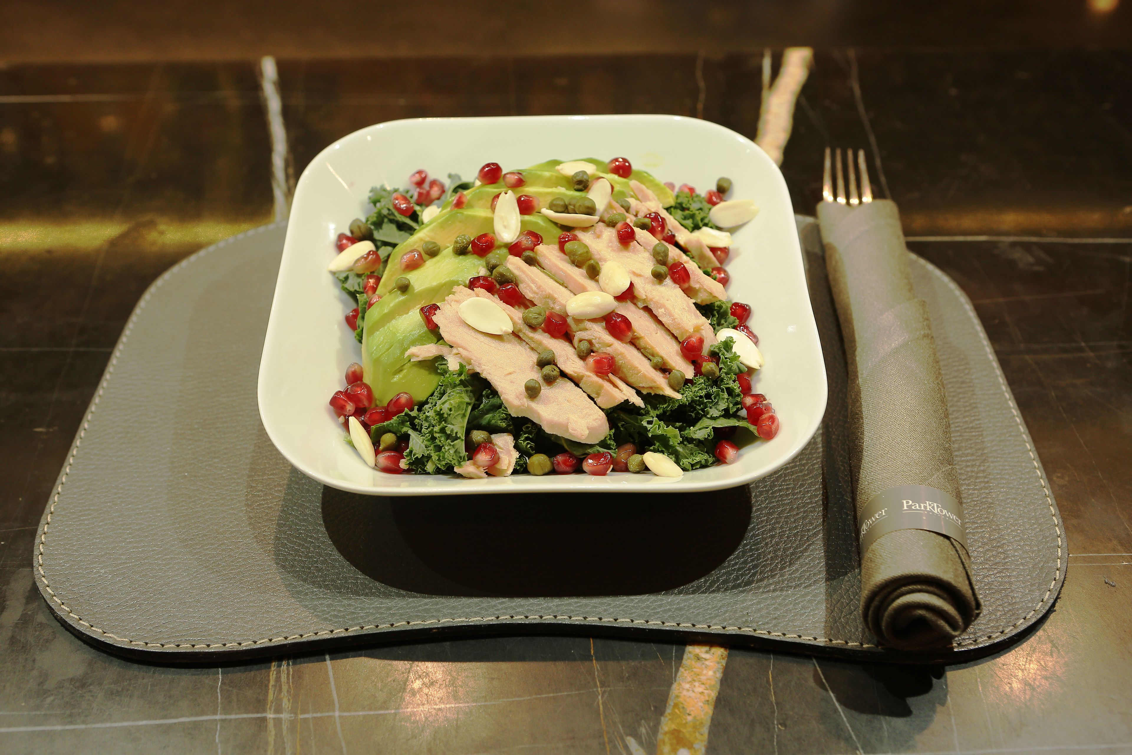 A white rectangular dish with sliced chicken, avocado, kale, and pomegranate seeds on a dark plate, placed on a textured table mat.