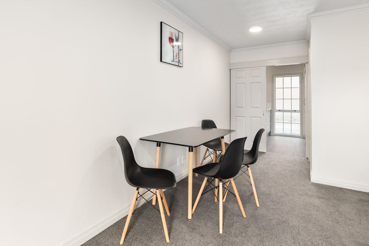 A room with a black table and four black chairs with wooden legs. There is a framed picture on the wall.