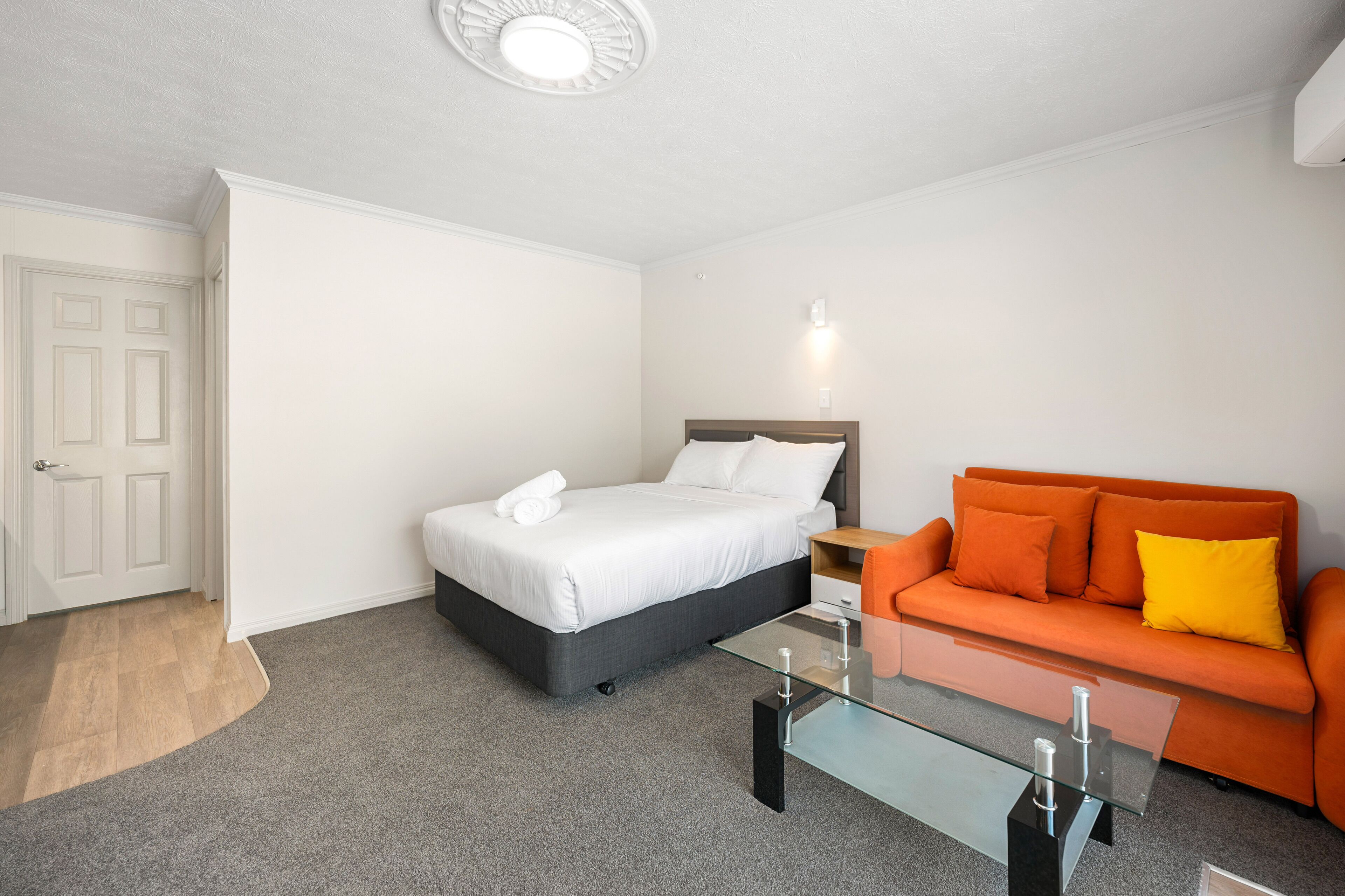 Un dormitorio con una cama, una mesita de centro de vidrio, un sofá naranja y una puerta blanca.