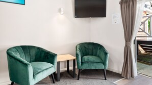 A room with two green armchairs, a small wooden table, a flat-screen TV mounted on the wall, and a window with curtains. - Ashleigh Court Motel (Rotorua)