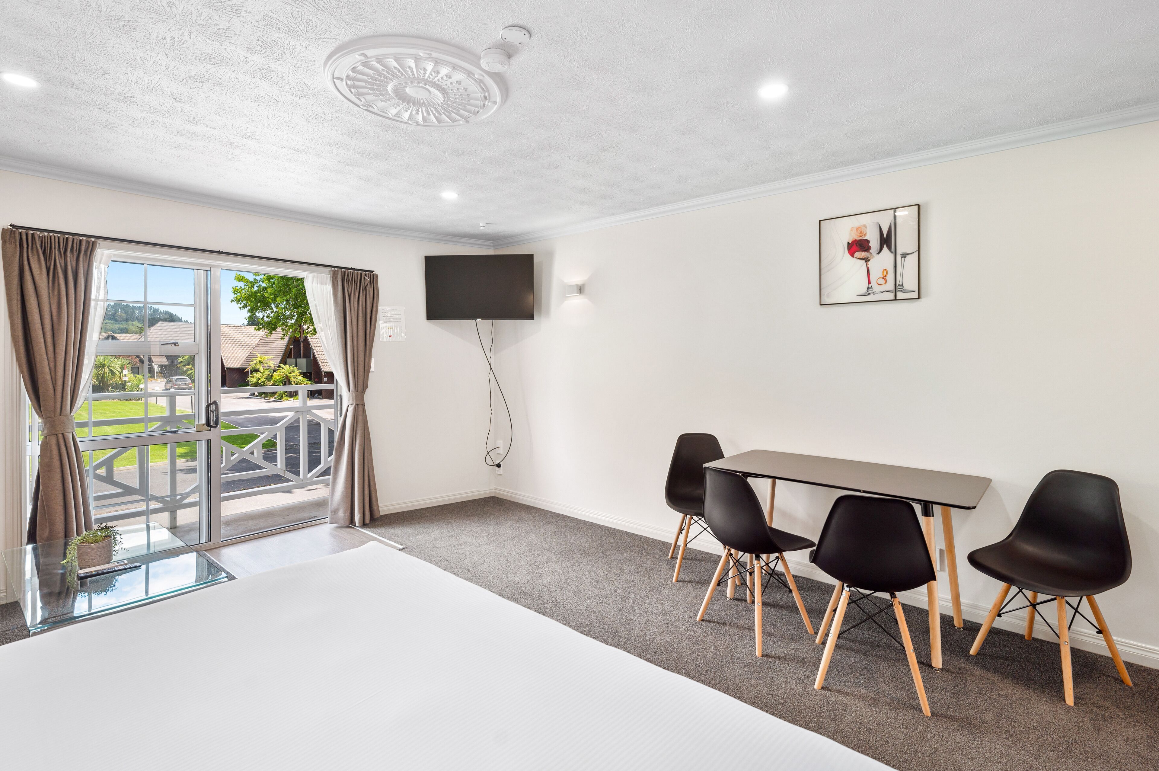 A modern room with a large window, a flat-screen TV, a dining table with four chairs, and a ceiling light fixture.