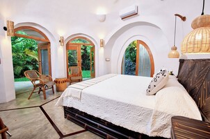 Un dormitorio con una cama grande, cabecera de madera y vistas a un follaje exuberante a través de puertas arqueadas.