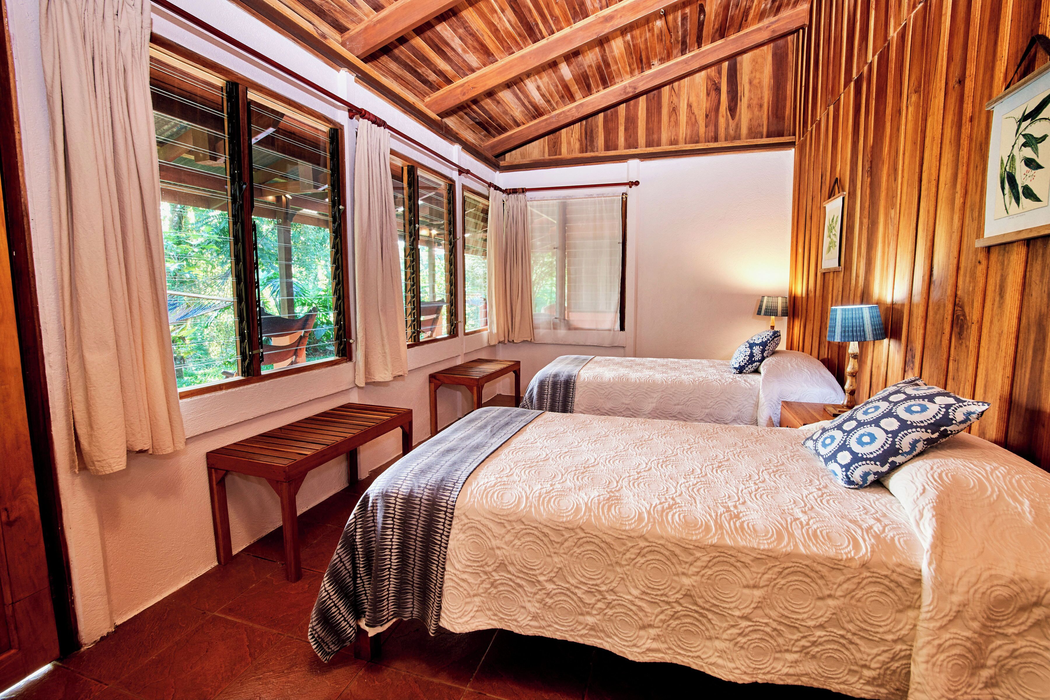 A room with two beds, wooden ceiling, large windows with curtains, a bench, and a small table.