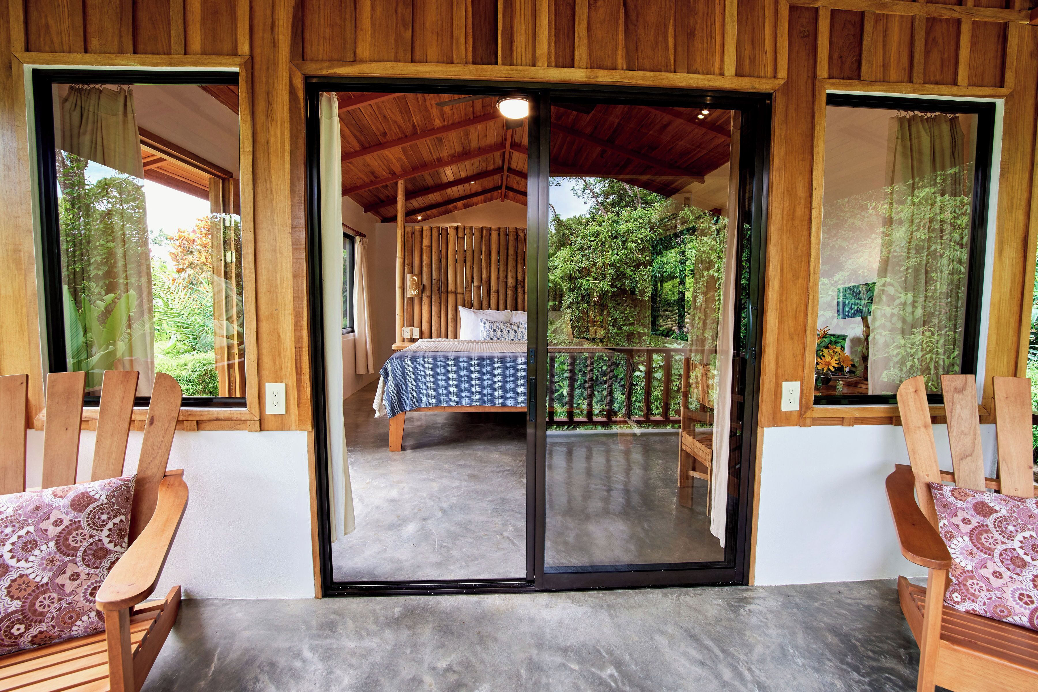 A wooden-framed room with a bed, a sliding glass door leading to a balcony, and two wooden chairs with patterned cushions.