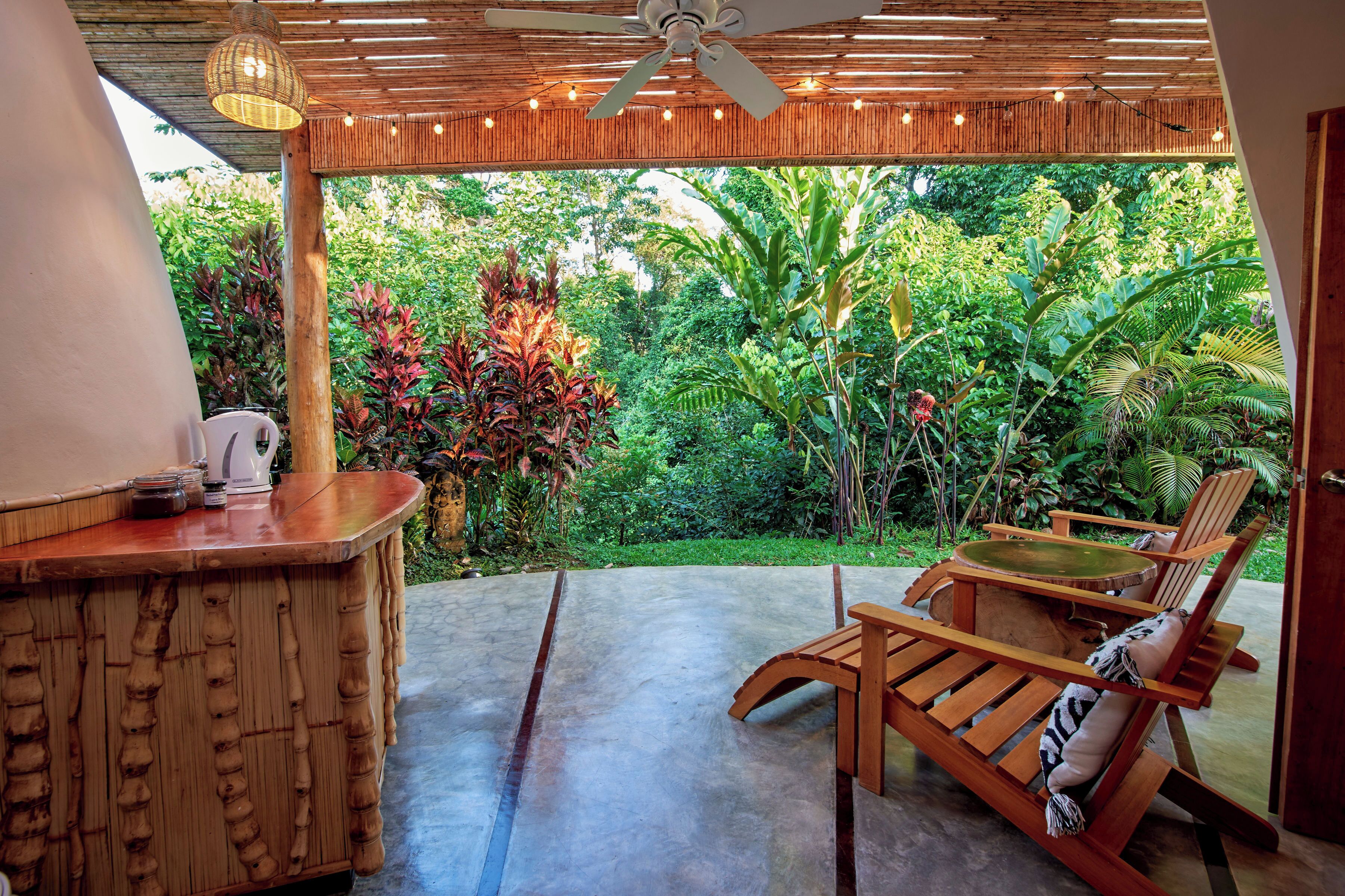 A wooden outdoor seating area with a table and chairs, a ceiling fan, and a view of lush greenery.