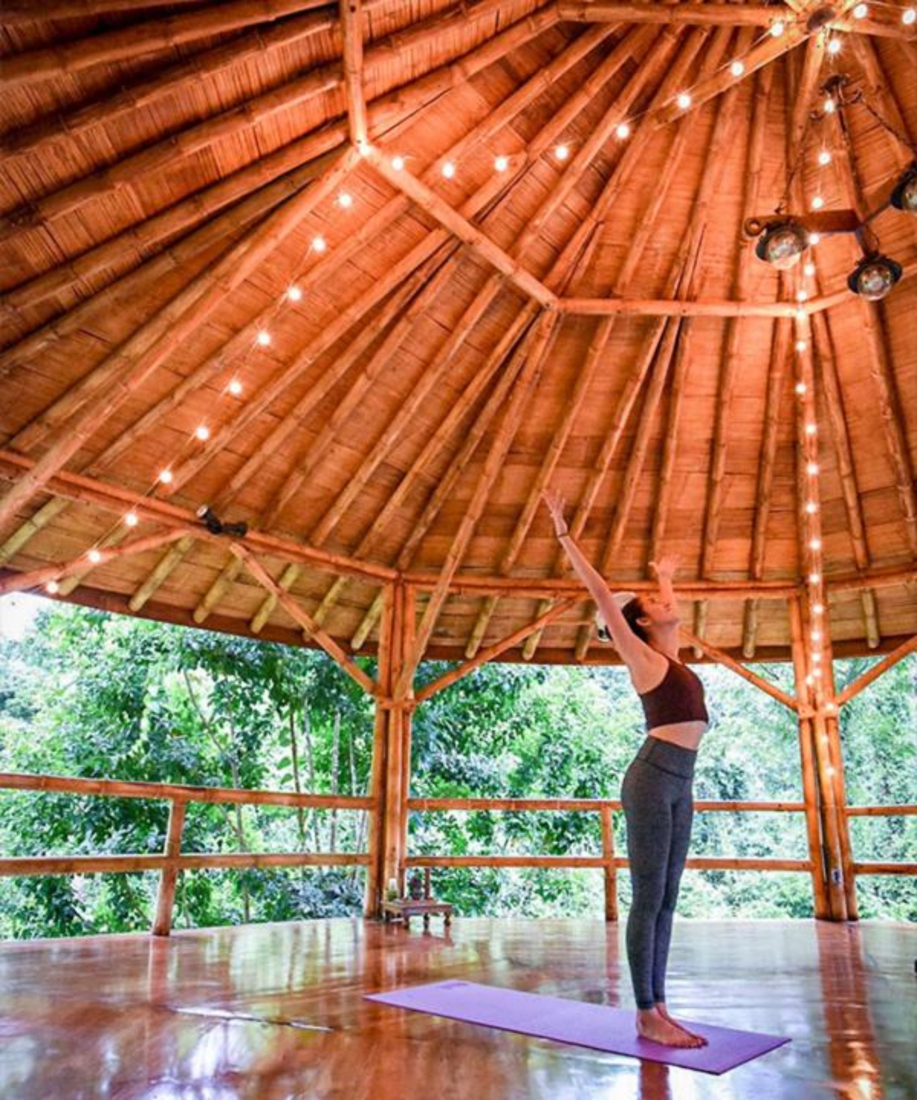 A person practicing yoga on a mat inside a wooden structure with a thatched roof and string lights.