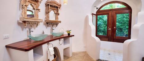 A bathroom with a wooden vanity, two ornate mirrors, a glass sink, and a view through an arched doorway.