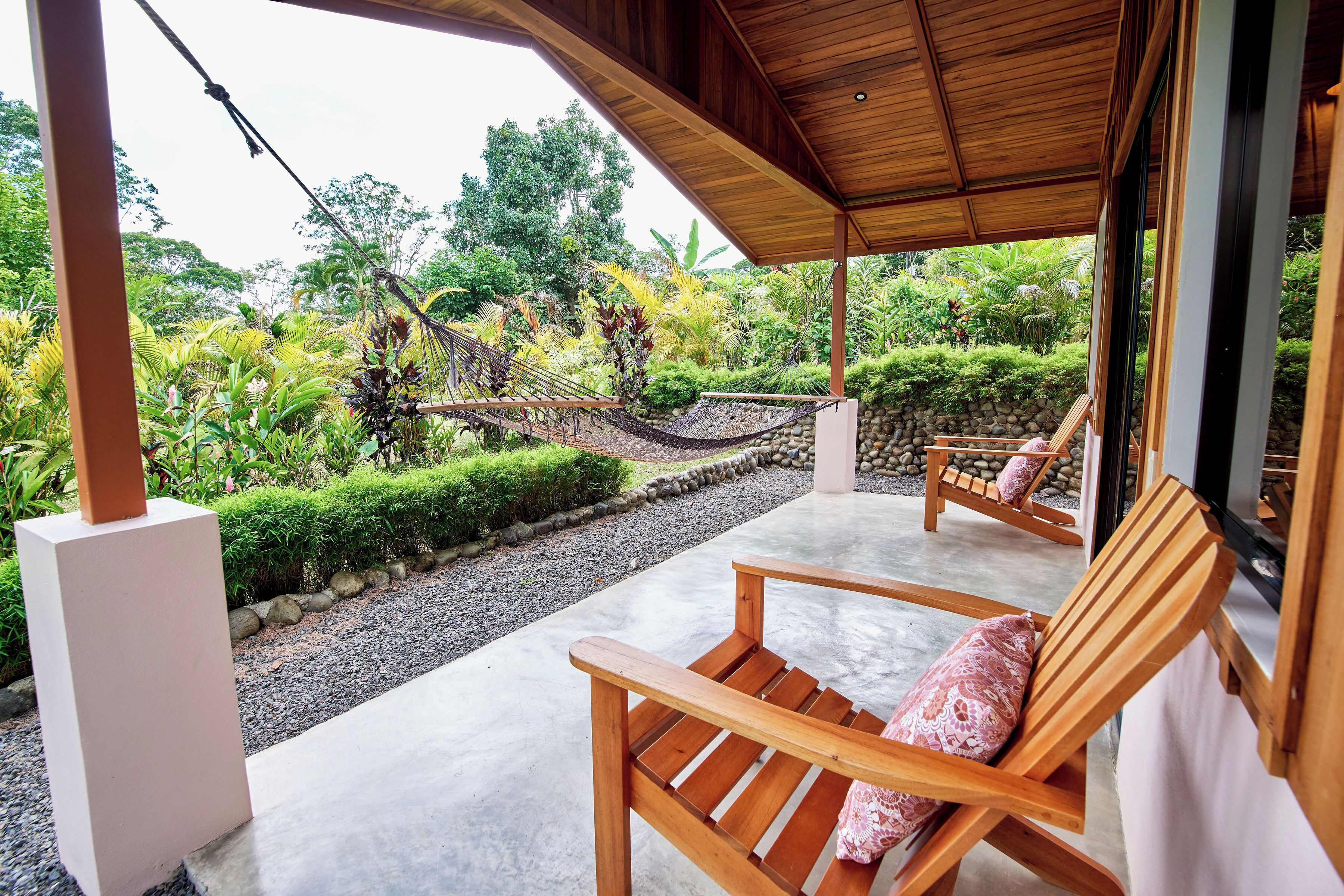 A wooden deck with two wooden chairs and a hammock, overlooking a lush garden with various plants and a stone pathway.