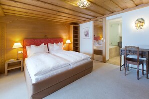 Quarto de hotel com uma cama grande, abajures, uma mesa com cadeira e um banheiro visível através de uma porta aberta.