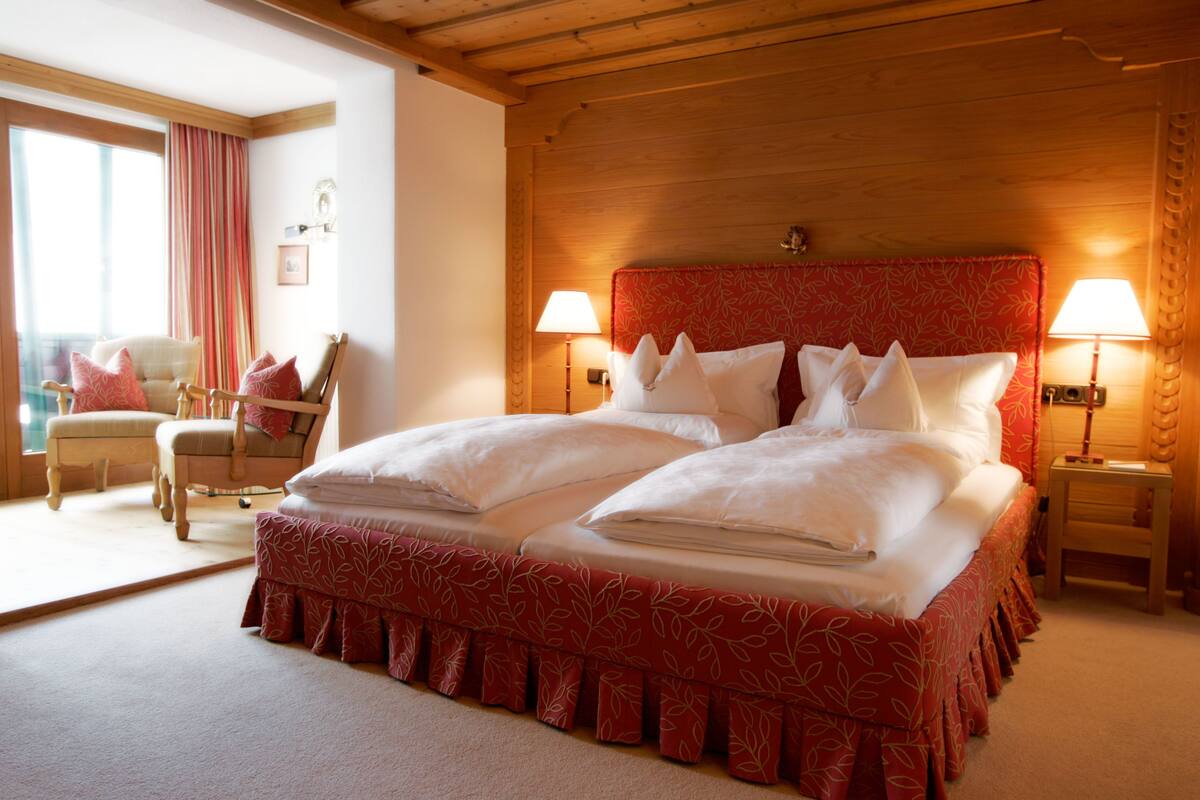 A double bed with a red headboard, two bedside tables with lamps, a wooden ceiling, and a window with curtains.
