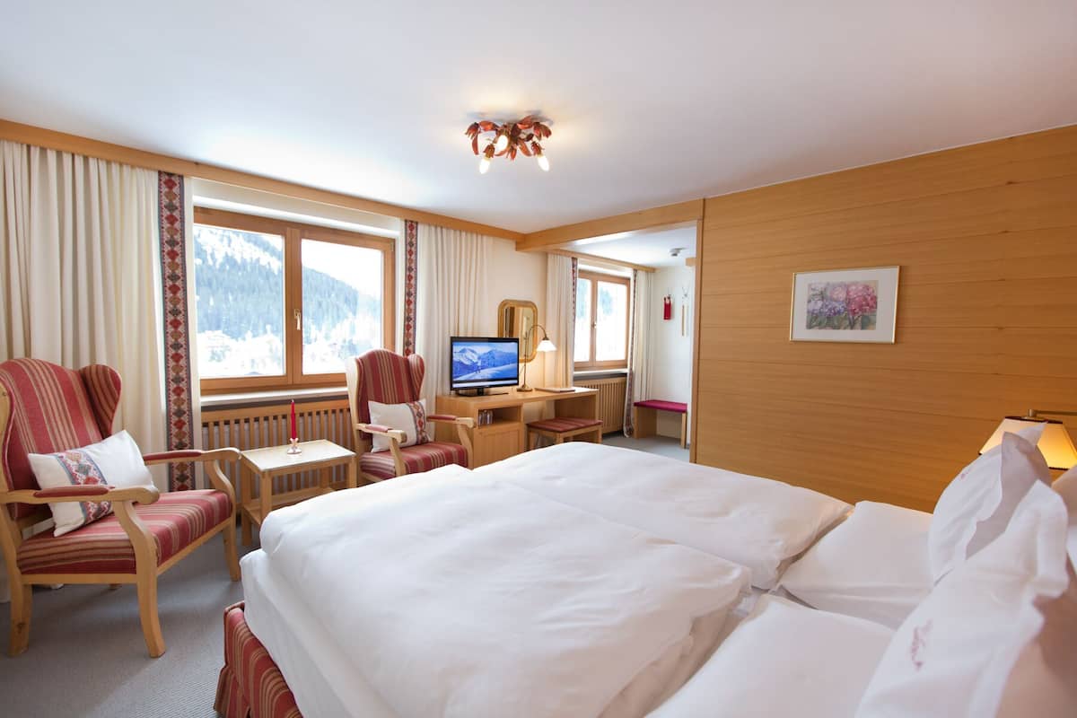A hotel room with a bed, two armchairs, a desk with a TV, and a window with a mountain view.